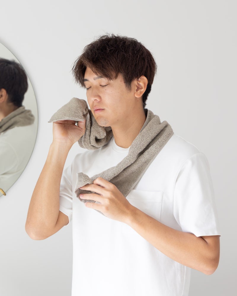トープカラーのちょうどいいマフラータオルを首にかけて洗顔後の顔を拭く男性。鏡のあるバスルームシーンで、柔らかな肌触りと日常使いの快適さを表現。