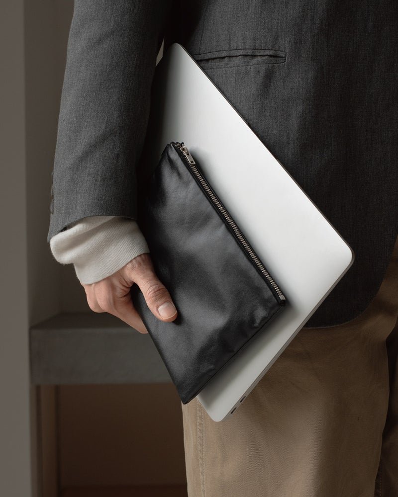 スーツ姿でFOLMAレザーポーチLとMacBookを一緒に手持ちするビジネスシーン。A4書類に近いLサイズがノートPCと並べて携帯できる実用的なクラッチ使いを提案した写真。