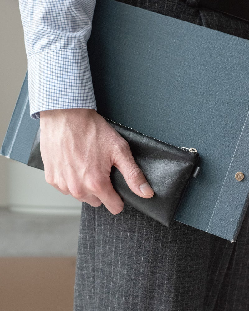 スーツ姿でFOLMAレザーポーチPENとクリップボードを一緒に手持ちするビジネスシーン。細長いPENサイズがバインダーに沿って収まりやすく会議や外出先でも上品に携帯できる。