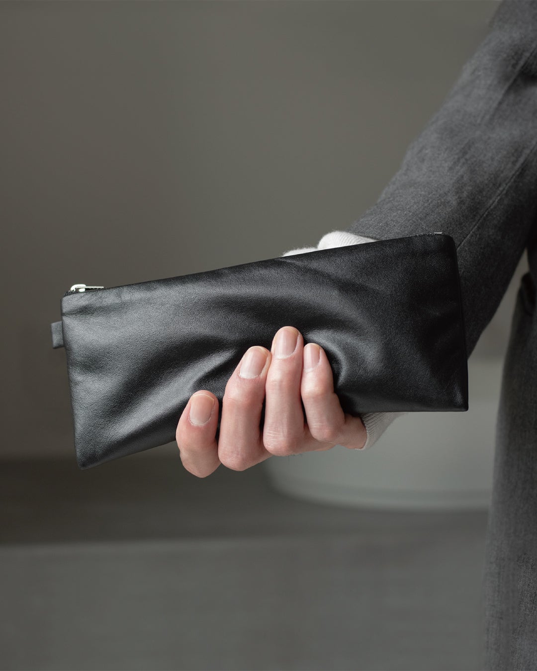 片手でFOLMAブラックナッパーレザーポーチPENを握り持つクローズアップ。200mmの細長フォルムが手にしっとりなじむ軽量本革ペンケースの持ちやすさを伝えるビジュアル。