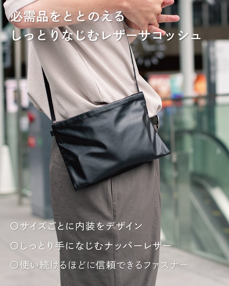 スーツ姿でFOLMAレザーサコッシュをショルダーストラップで斜めがけして手を添えるビジネスコーデ。ナッパーレザーとYKKファスナー採用で毎日の必需品をしっとりなじむ本革でまとめる。