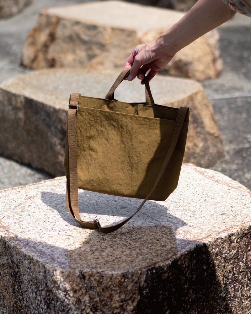石の上に置かれたブラウンの2way Pocket TOTEを手に持ち上げる場面で、コンパクトなサイズ感と自然な素材の質感が伝わるカット。
