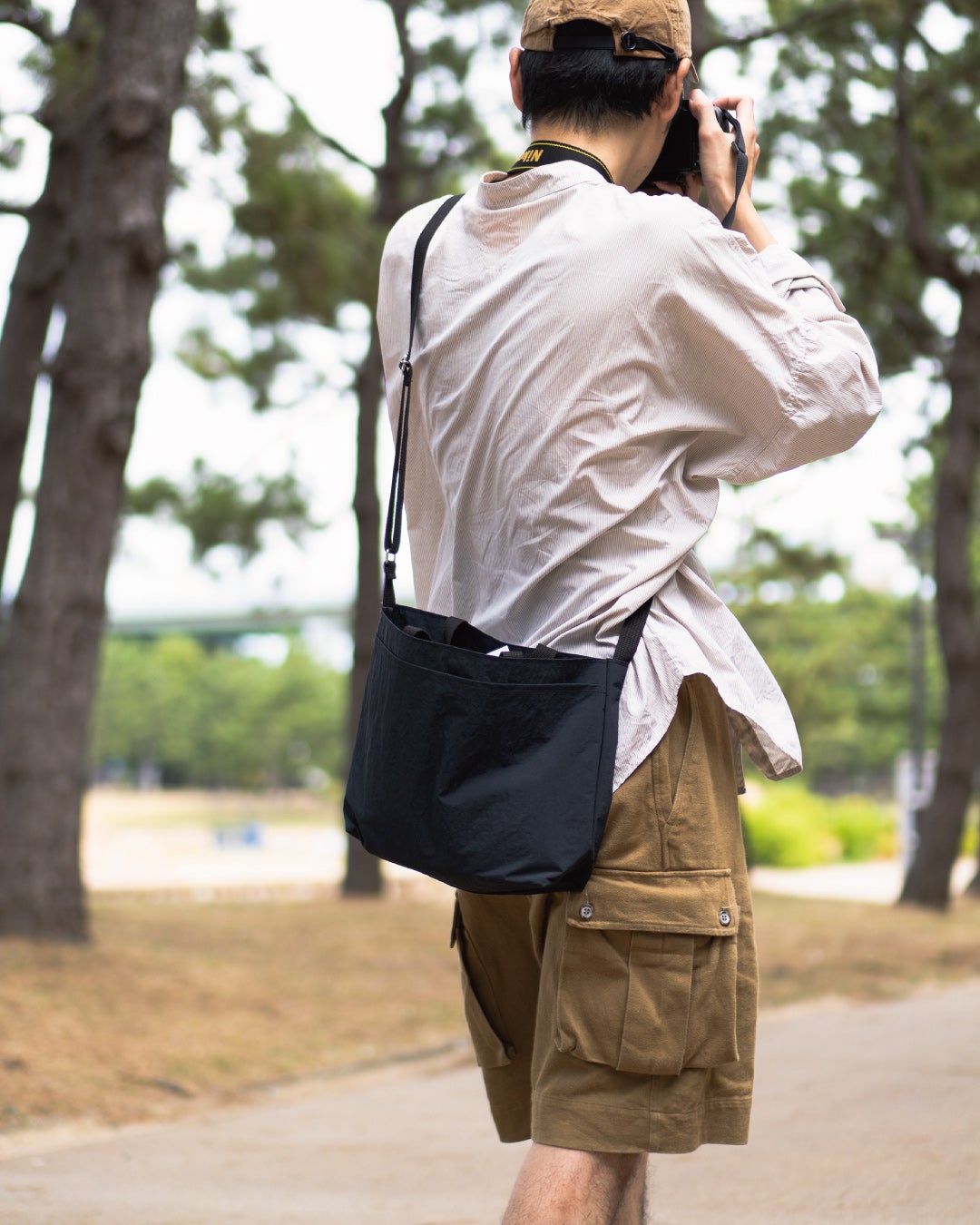 黒の2way Pocket TOTEを斜め掛けしてカメラを構える男性の後ろ姿で、写真撮影など外出時に活躍するクロスボディスタイルを示す一枚。