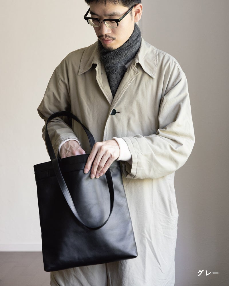 レザートートの中を確かめる男性が身につけた、グレーの3秒マフラー キッドモヘヤとベージュコートの軽やかな重ね着をとらえた日常のワンシーン。